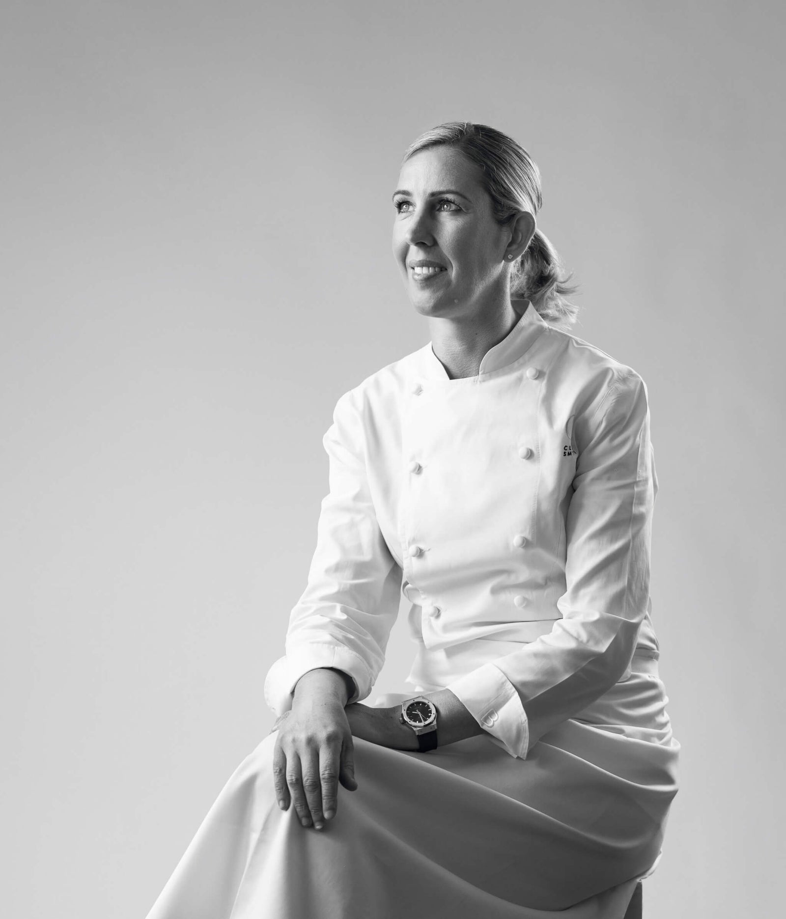 female chef in a white chef coat sitting with a thoughtful expression in a minimalist background