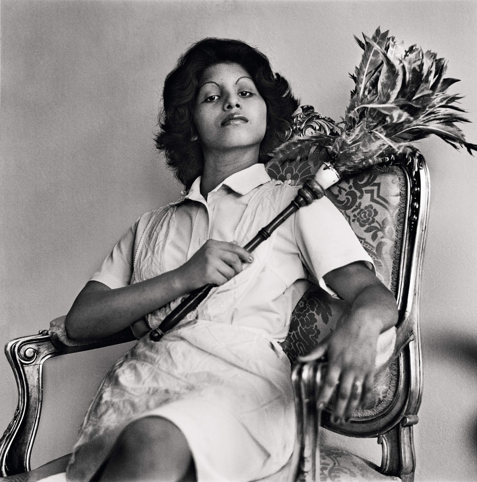 Young woman seated in an ornate chair, holding a decorative staff with a pineapple top, wearing a white shirt and apron, in a black and white photograph.