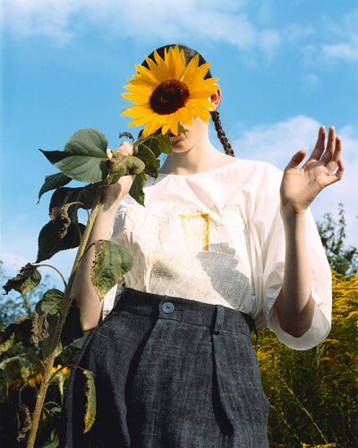 person holding a sunflower in front of their face, wearing a white blouse and dark trousers, surrounded by a garden with blue sky and greenery