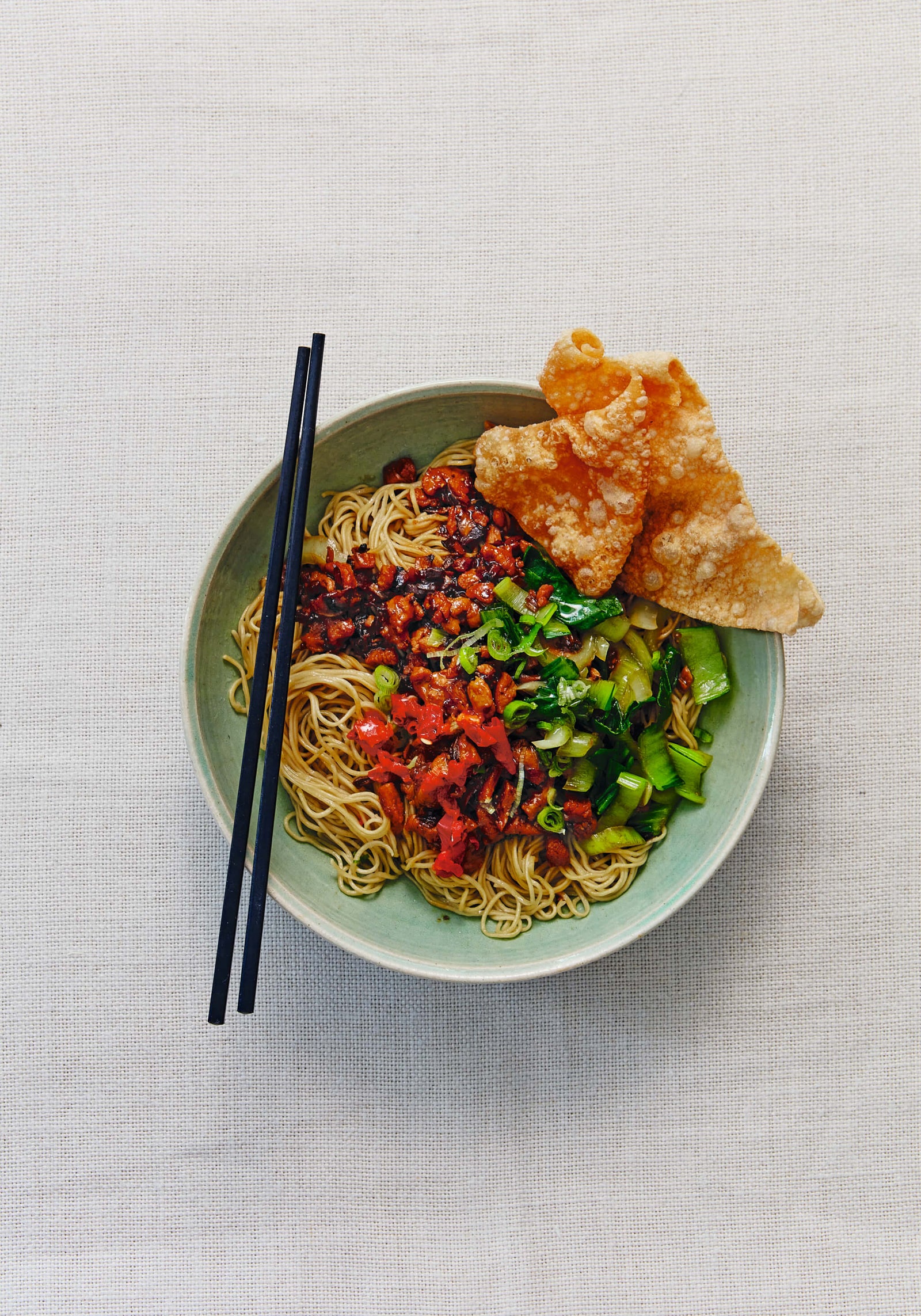bowl of noodles topped with chopped vegetables, spicy sauce, and crispy fried wontons, served with black chopsticks on a light linen background