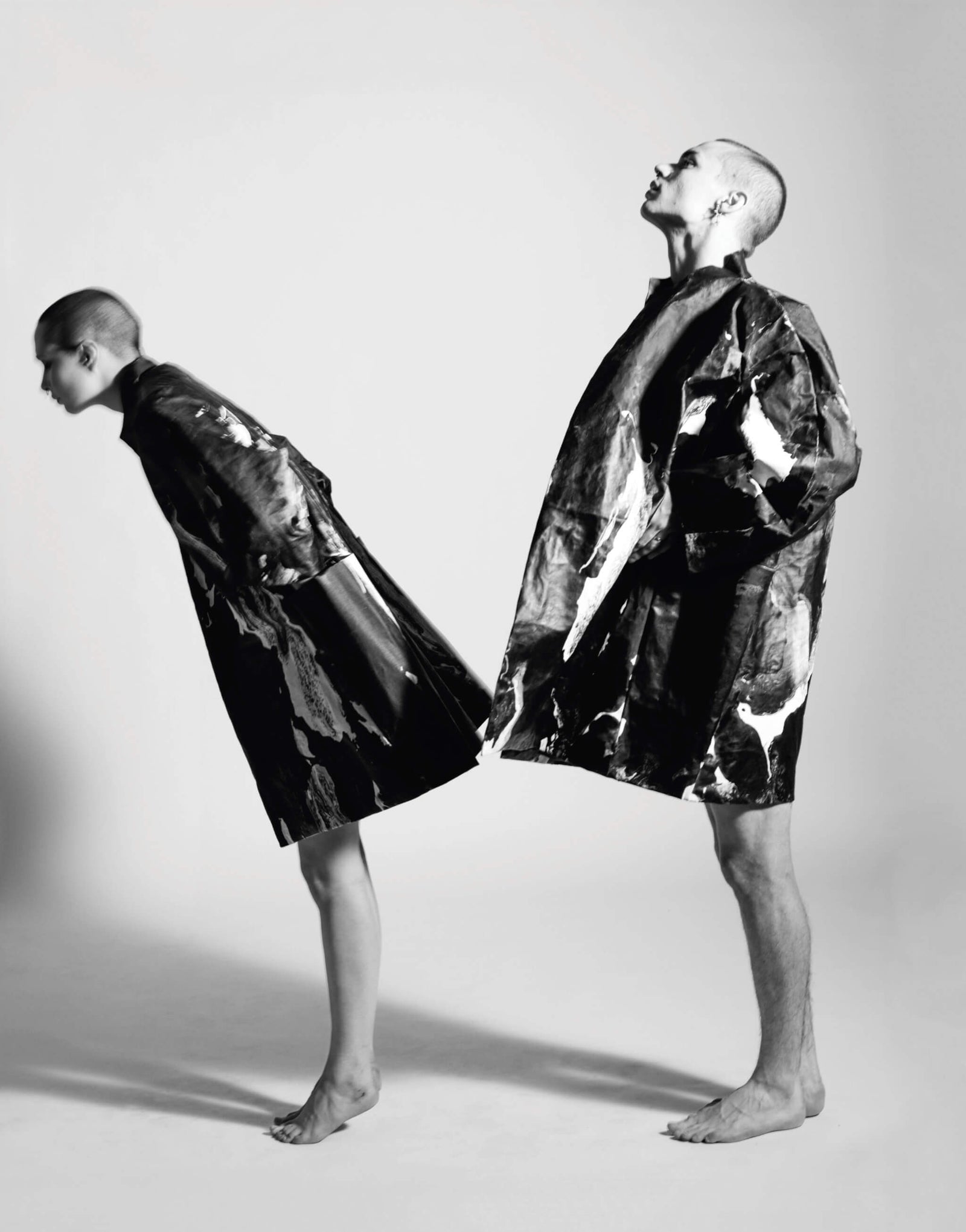 two models in black and white wearing abstract-patterned oversized coats, one model leaning forward and the other standing upright, minimalist fashion photography