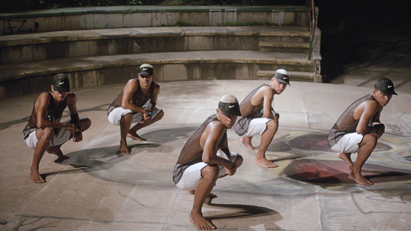 dance performance at night featuring four dancers in matching attire, squatting in a semi-circle on a graffiti-covered surface, wearing black Nike hats and sleeveless shirts, barefoot, showcasing hip-hop culture and street style