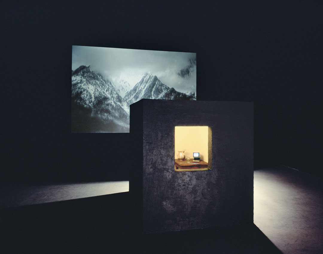 Bill Viola, Room for St. John of the Cross, 1983.Video and sound installation in a dark room, with a black cubicle with window, peat moss, wooden table, glass and metal pitcher with water, color video, and one-channel mono sound; black-and-white video projection; amplified stereo sound; room: 14 x 24 x 30 ft. (4.3 x 7.3 x 9.1 m); projected image: 8 ft. 7 in. x 12 ft. 8 in. (2.6 x 3.7 m); continuously running. Photo: Kira Perov