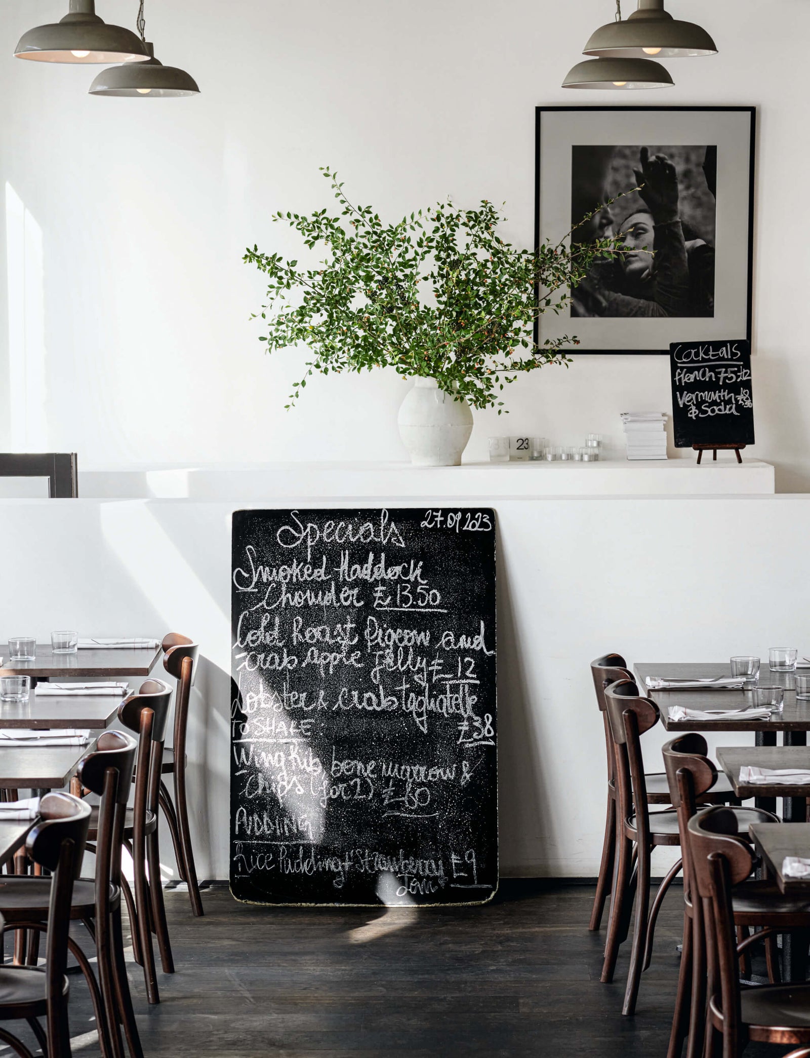 interior of a modern restaurant featuring a chalkboard menu with daily specials, a decorative plant in a vase, wooden tables and chairs, and pendant lighting