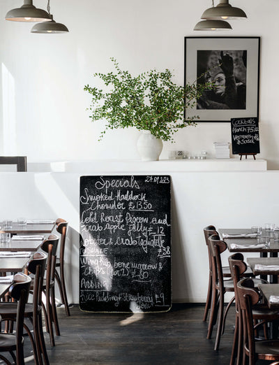 interior of a modern restaurant featuring a chalkboard menu with daily specials, a decorative plant in a vase, wooden tables and chairs, and pendant lighting