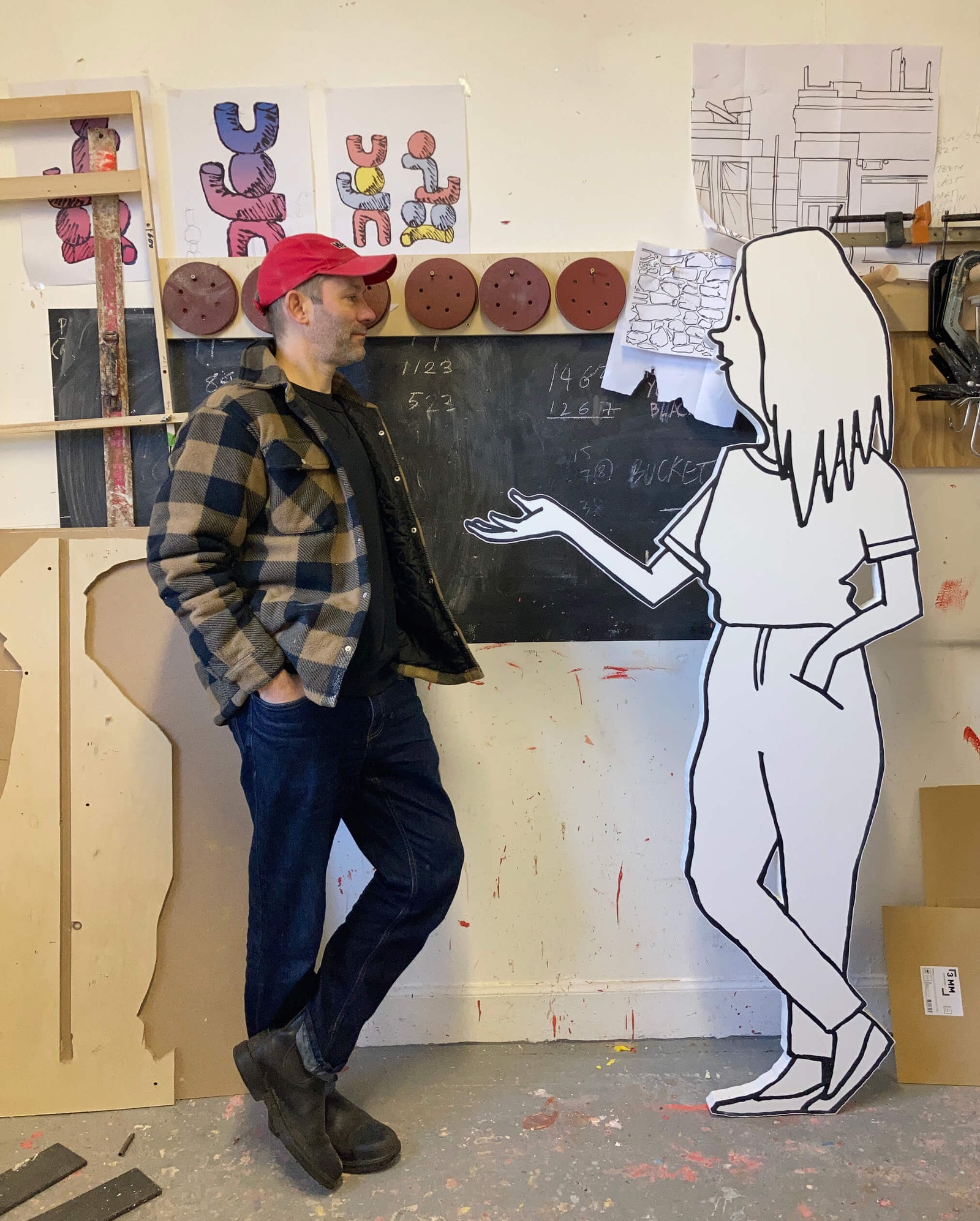 man in a plaid shirt and red hat standing next to a white illustrated figure in a creative studio with colorful wall art and wood materials