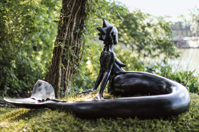 sculpture of a mermaid sitting on the grass surrounded by greenery and a tree in the background