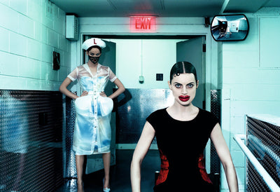 two fashion models in a surreal setting, one wearing a transparent nurse dress and cap, the other with bold red lips and a black dress, industrial background with an EXIT sign and security mirror