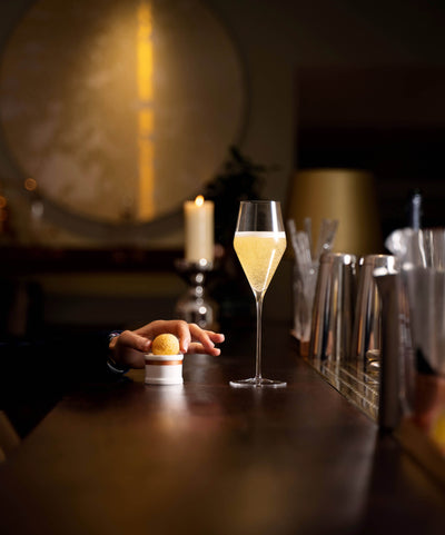 hand reaching for a small dish with a golden ball-shaped appetizer next to a tall glass of champagne on a wooden bar top in a dimly lit setting with a candle and decorative background elements