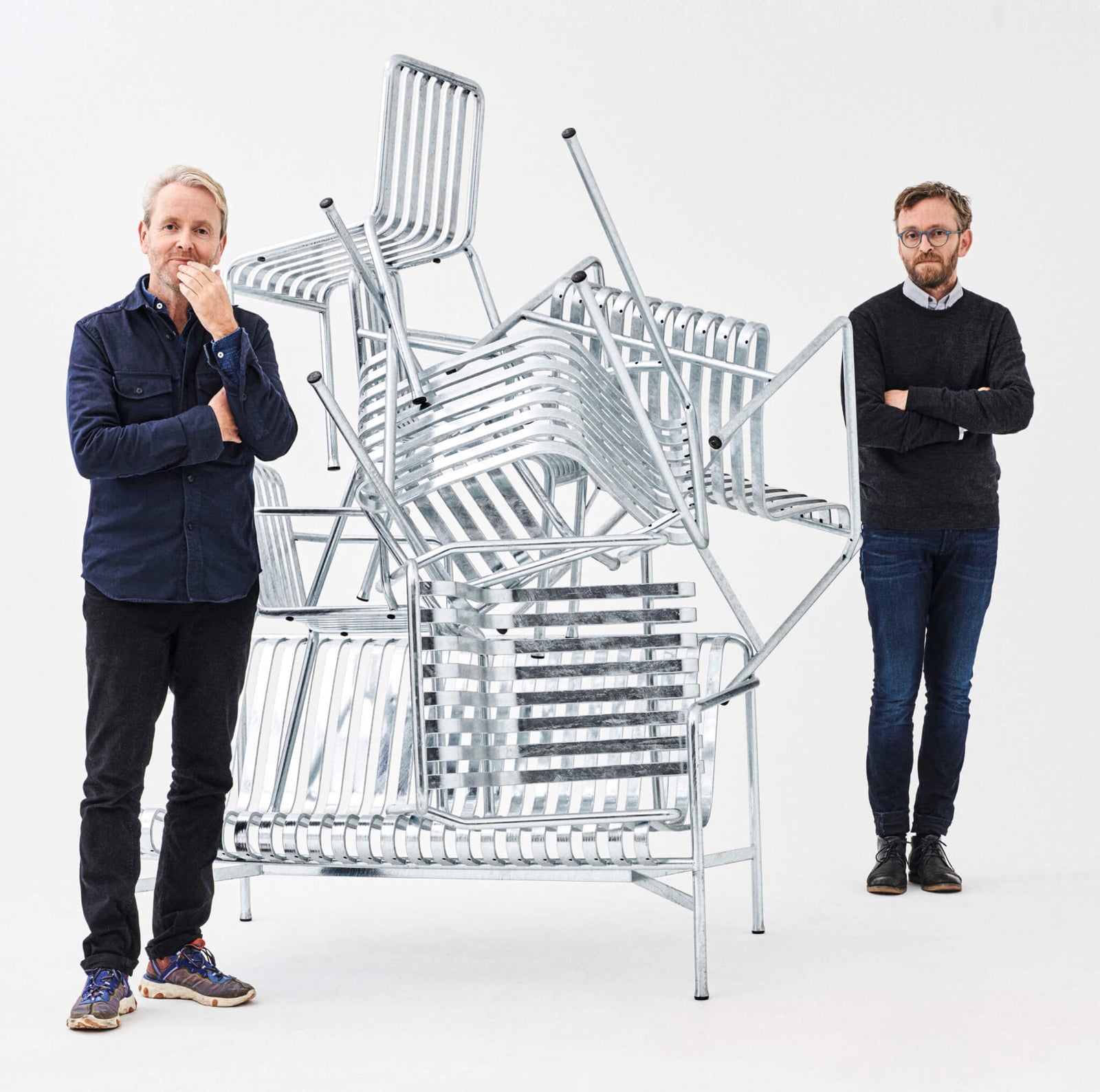 two men standing beside a stack of metal chairs against a white background, one man is touching his chin thoughtfully, while the other has his arms crossed, focusing on the chair arrangement
