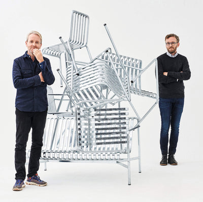 two men standing beside a stack of metal chairs against a white background, one man is touching his chin thoughtfully, while the other has his arms crossed, focusing on the chair arrangement