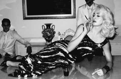 woman in elegant black and white dress smoking a cigarette, lying on a table surrounded by two men in formal attire, vintage setting with ornate decor and elegant furnishings