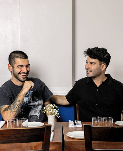 Two smiling men enjoying a meal in a restaurant, seated at a wooden table with white plates, glassware, and a small flower vase, creating a warm dining atmosphere.