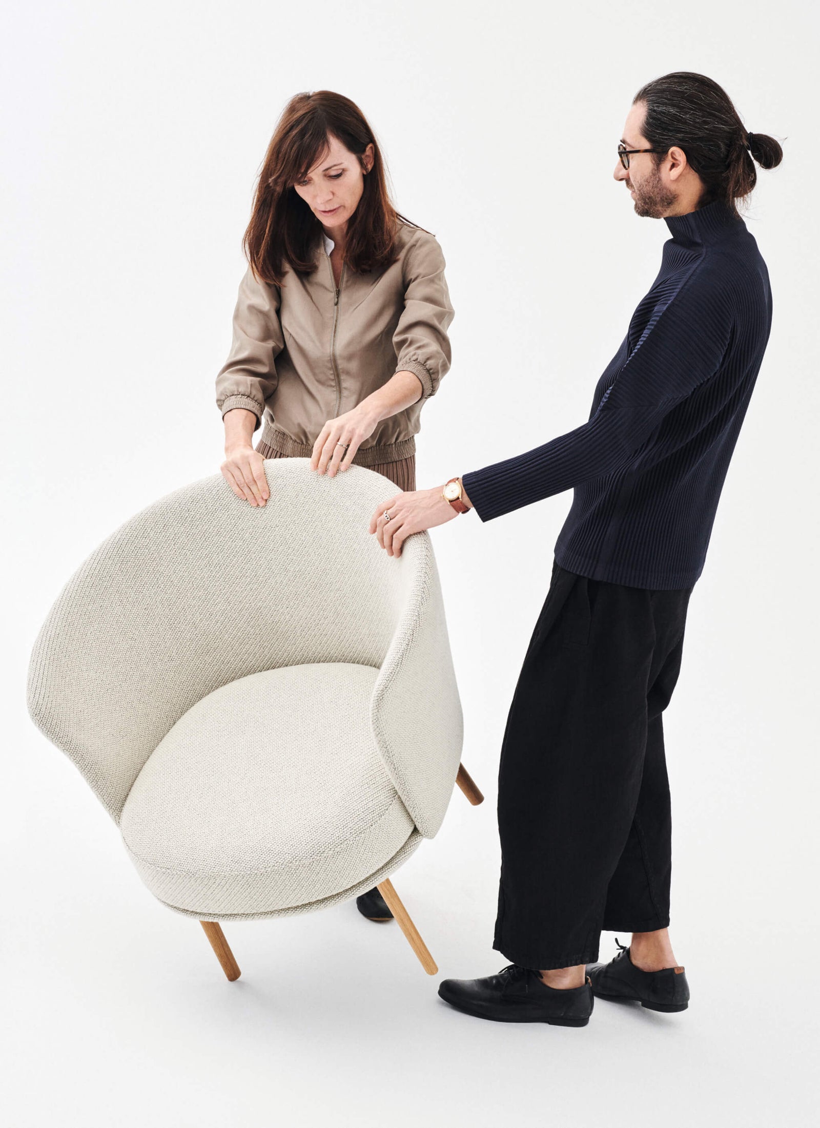 two people assembling a modern cream-colored armchair with wooden legs in a minimalist studio setting