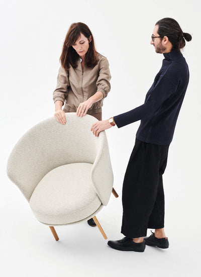 two people assembling a modern cream-colored armchair with wooden legs in a minimalist studio setting