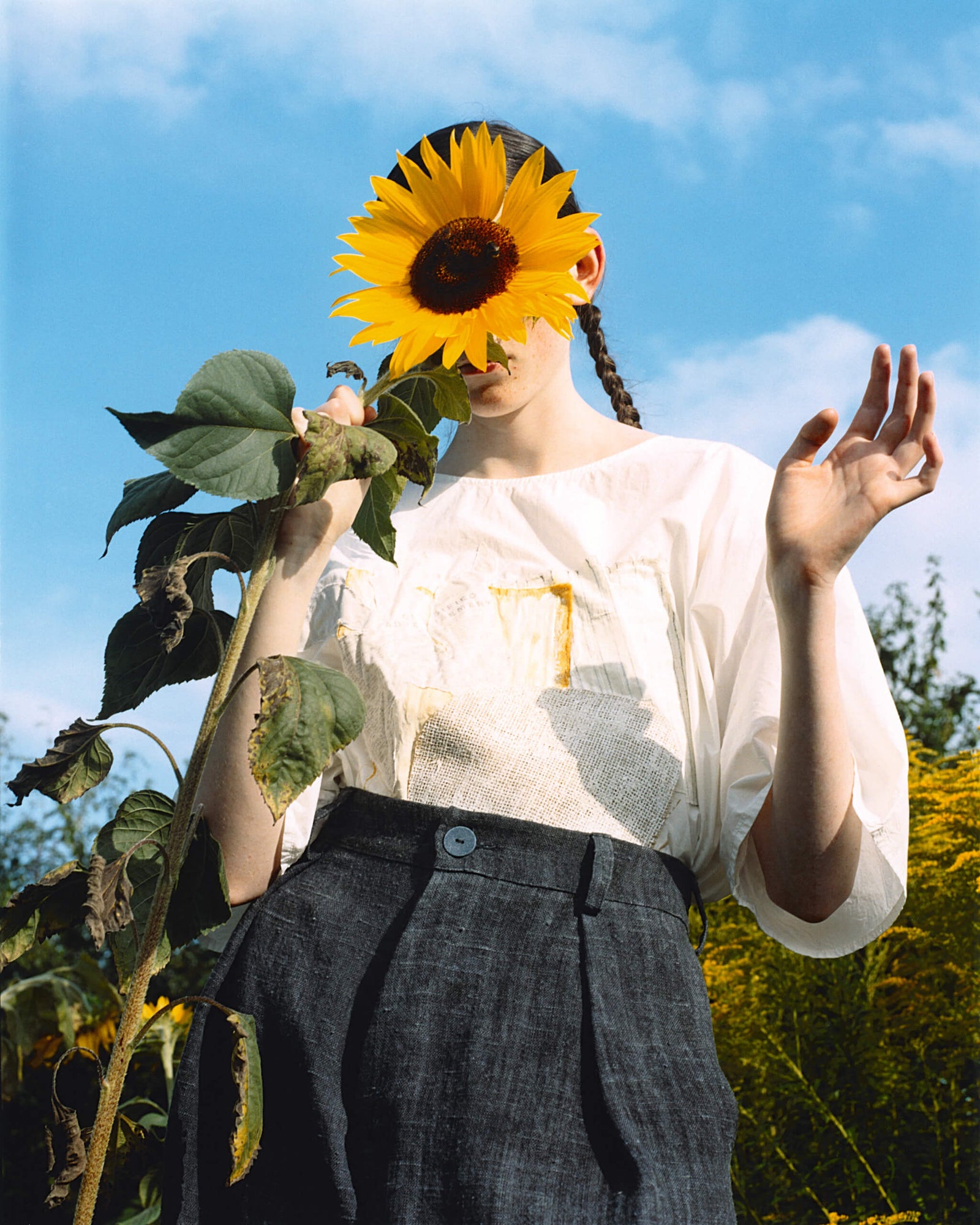 person holding a sunflower in front of their face, wearing a white blouse and dark trousers, surrounded by a garden with blue sky and greenery