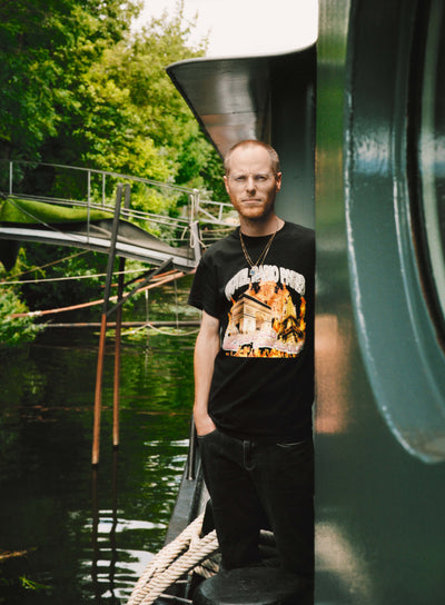 Man with glasses standing on a boat next to a green wall, wearing a black t-shirt with a graphic design, surrounded by greenery and water, in a calm outdoor setting.
