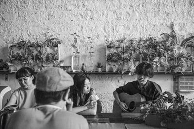 group of people sitting in a cozy café with plants on the wall, one person playing guitar, black and white photo