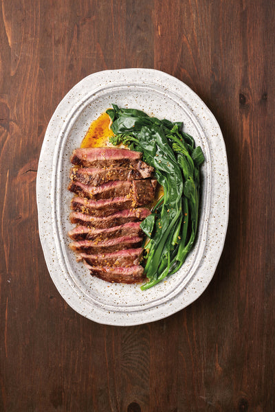 sliced beef steak served with sautéed greens on a white oval plate with a wooden table background
