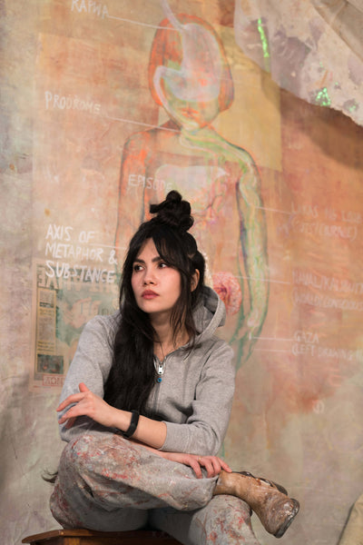 Young woman in a gray hoodie and paint-splattered pants sitting on a stool, reflecting in an art studio with a colorful mural in the background featuring abstract shapes and text.