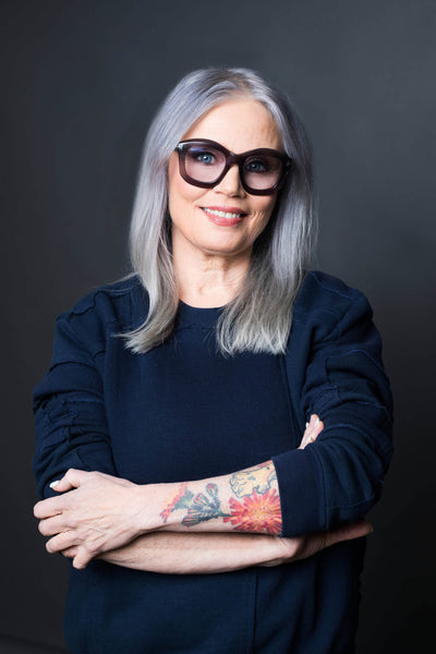 portrait of a smiling woman with long gray hair, wearing oversized sunglasses and a navy blue sweater, showcasing colorful floral tattoos on her arms against a dark background