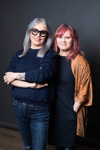 Two women posing together for a portrait, one with long gray hair and glasses wearing a dark sweater, the other with medium-length pink hair in a brown patterned shawl, against a dark background.