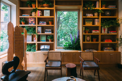 cozy reading nook with wooden shelves, books, potted plants, and two chairs by a large window showcasing greenery outside