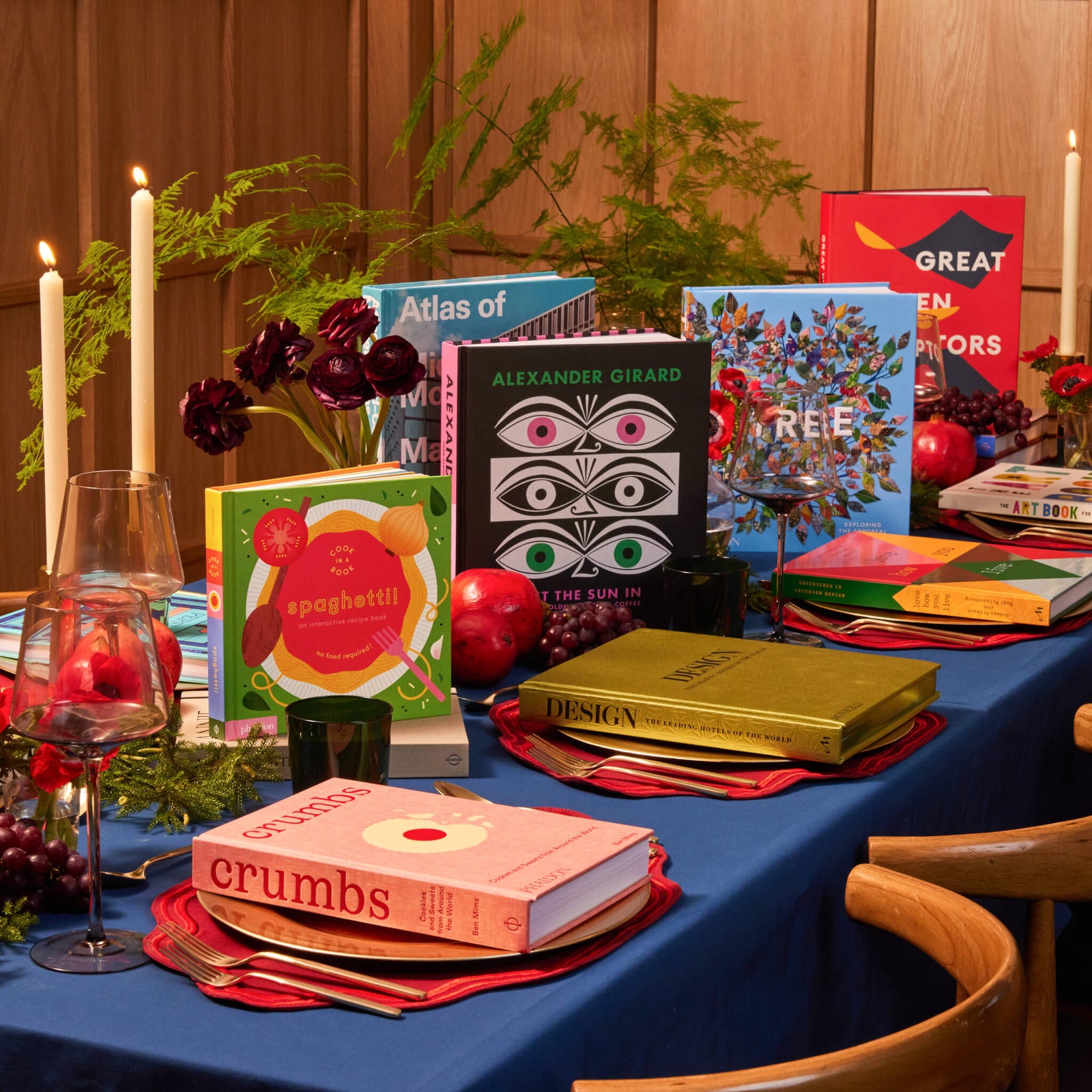 a beautifully arranged dining table featuring colorful books on design, cooking, and art, surrounded by decorative elements like fruit, candles, and greenery, creating a cozy and inviting atmosphere for a gathering