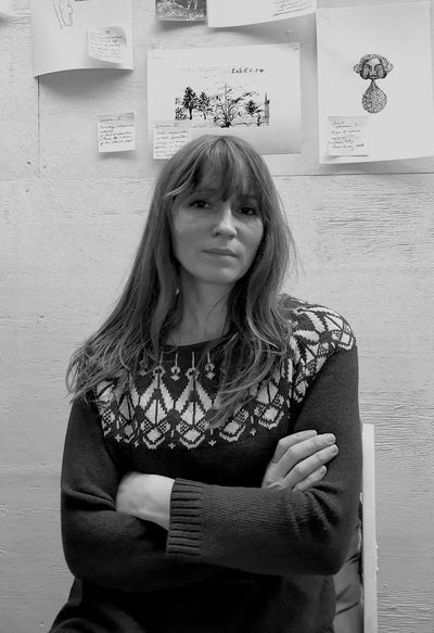 Woman with long hair wearing a patterned sweater, seated with arms crossed, in front of a wall covered with sketches and notes in black and white.