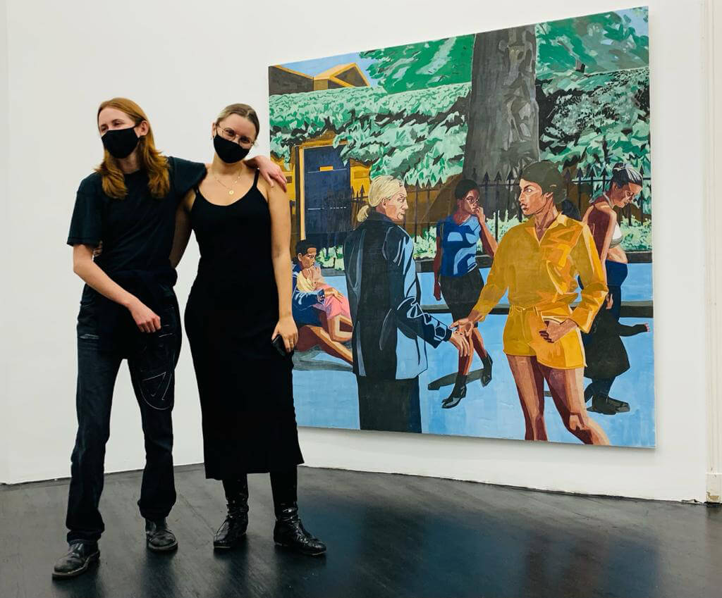 Two individuals posing in front of a colorful painting featuring diverse figures in a park setting, wearing black masks, showcasing a contemporary art gallery environment.