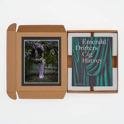 Photograph of a woman in a lavender gown under blooming wisteria, accompanied by the book "Emerald Drifters" by Cig Harvey in a cardboard box packaging.