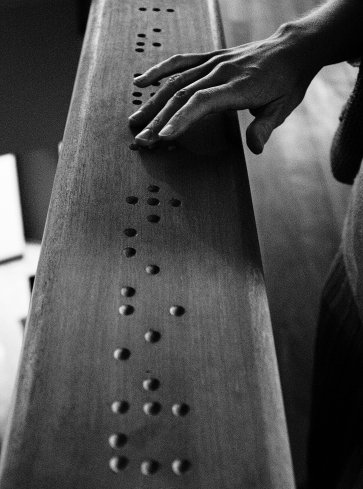 A hand reading wood-carved braille code where the word ⠏⠗⠑⠍⠊⠑⠗ (premier, French for 