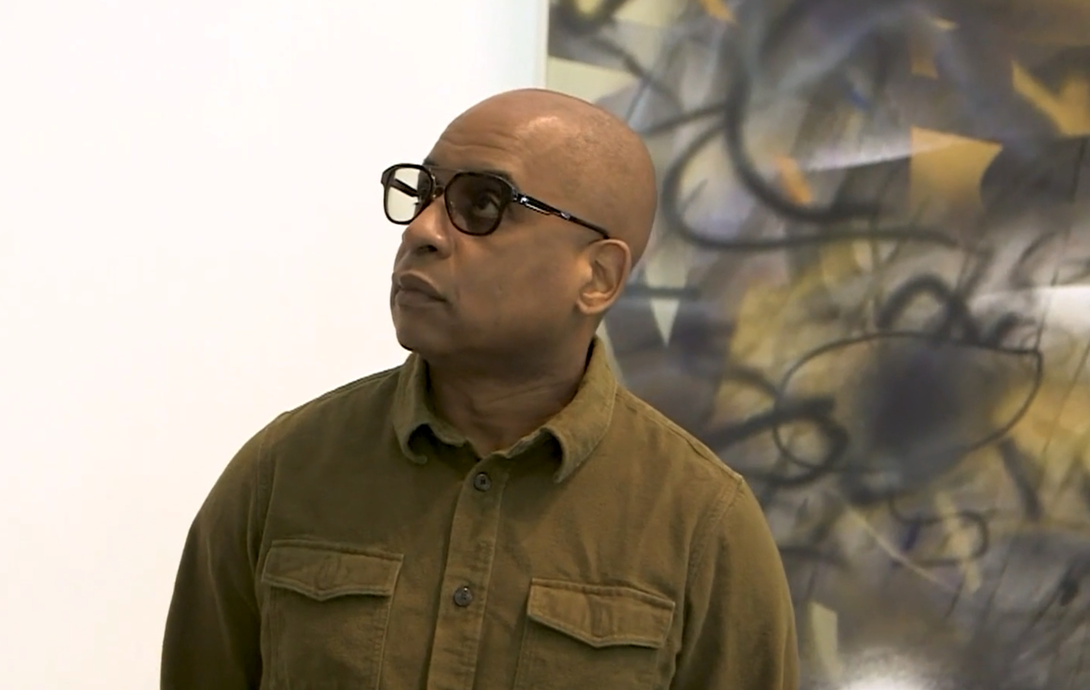 Glenn Ligon on PBS NewsHour at Grief and Grievance, at the New Museum, New York, beside a painting by Julie Mehretu